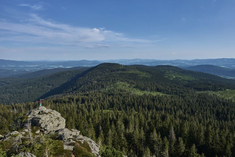 Wanderer stehen unter einem klaren blauen Himmel auf einem Felsvorsprung und blicken auf ein weites bewaldetes Tal und die majestätischen Berge bei Bodenmais.