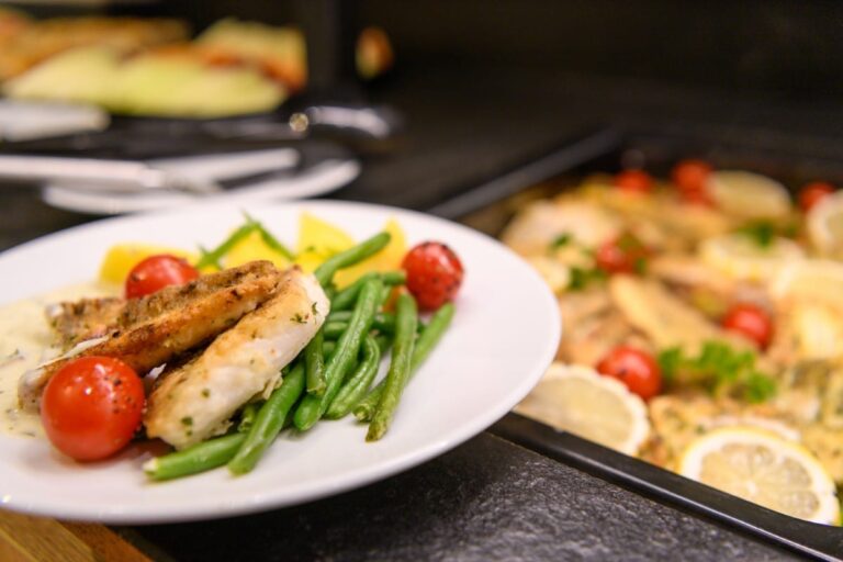 Ein Teller mit gegrilltem Fisch, grünen Bohnen, Kirschtomaten und Zitronenscheiben lädt in unserem Hotel Bodenmais zum Verweilen ein, neben ihm eine Schale mit gebackenem Fisch mit Zitrone.