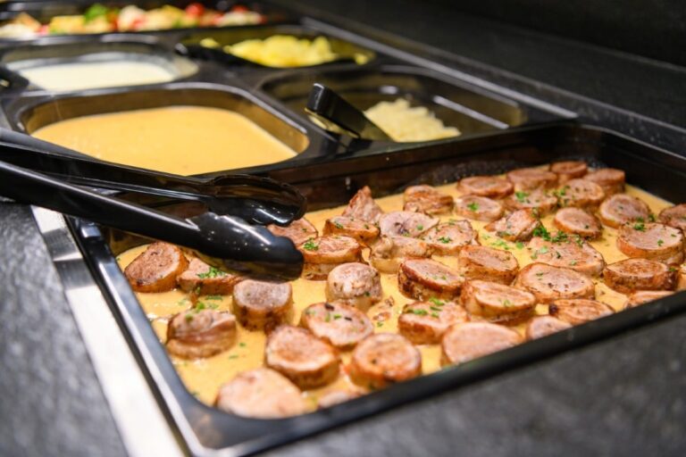Buffet-Tablett mit Wurstscheiben in pikanter Sauce, eingebettet neben einer Reihe anderer Gerichte im Hotel Bodenmais, Servierzangen stehen zum Einsatz bereit.