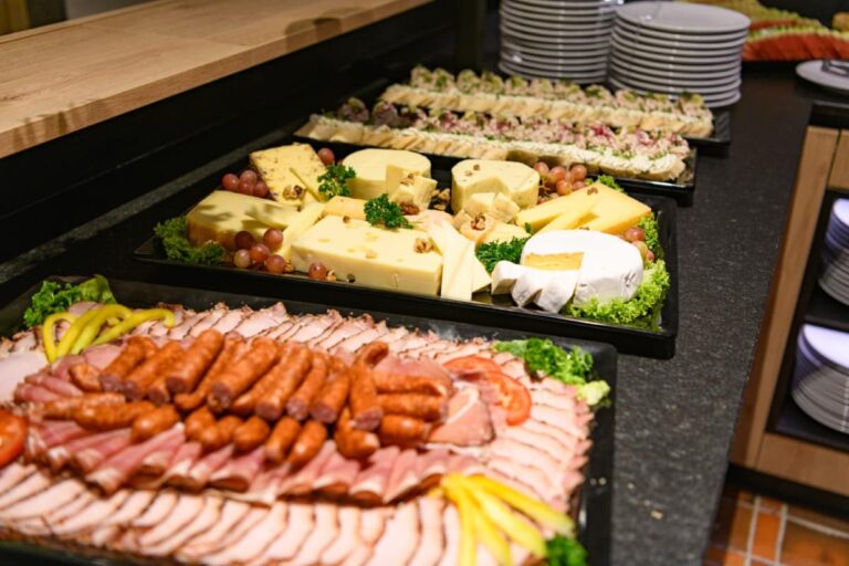 Im Hotel Bodenmais erwartet die Gäste eine elegante Auswahl an Wurst, Käse und Salaten. Im Hintergrund laden ordentlich gestapelte Teller zum Genießen ein.