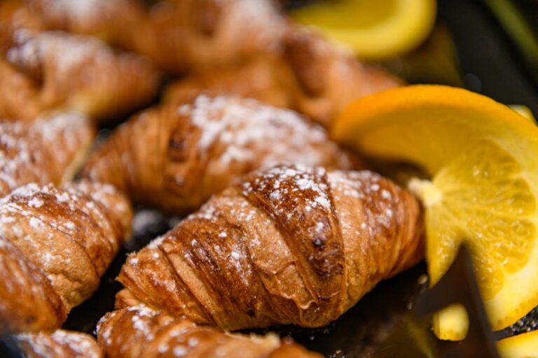 Eine Nahaufnahme von blättrigen Croissants, bestäubt mit Puderzucker und garniert mit leuchtend orangefarbenen Scheiben, erinnert an ein herrliches Frühstück im Hotel Bodenmais.