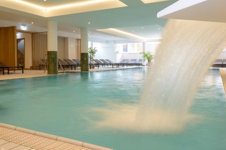 Entspannen Sie im Hotel Bodenmais am Innenpool mit Liegestühlen, wo das Wasser sanft aus einem Ausguss auf der rechten Seite herabstürzt. Das moderne Design wird durch sanfte Beleuchtung ergänzt und schafft eine ruhige Atmosphäre.