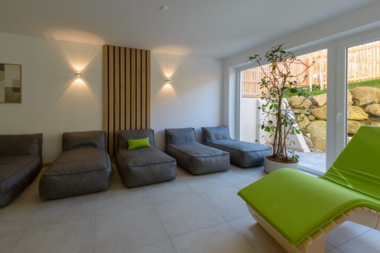 Erleben Sie die ruhige Eleganz eines minimalistischen Zimmers im Hotel Bodenmais mit grauen Loungesesseln, einem grünen Kissen, einer Topfpflanze und einer hellgrünen Liege neben großen Fenstern, die natürliches Licht hereinlassen.