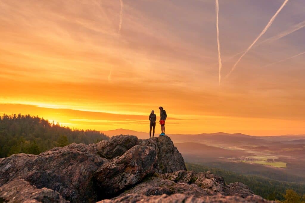 Zwei Menschen stehen auf felsigen Klippen und blicken auf einen Wald bei Sonnenuntergang mit orangefarbenem Himmel und fernen Wegen.
