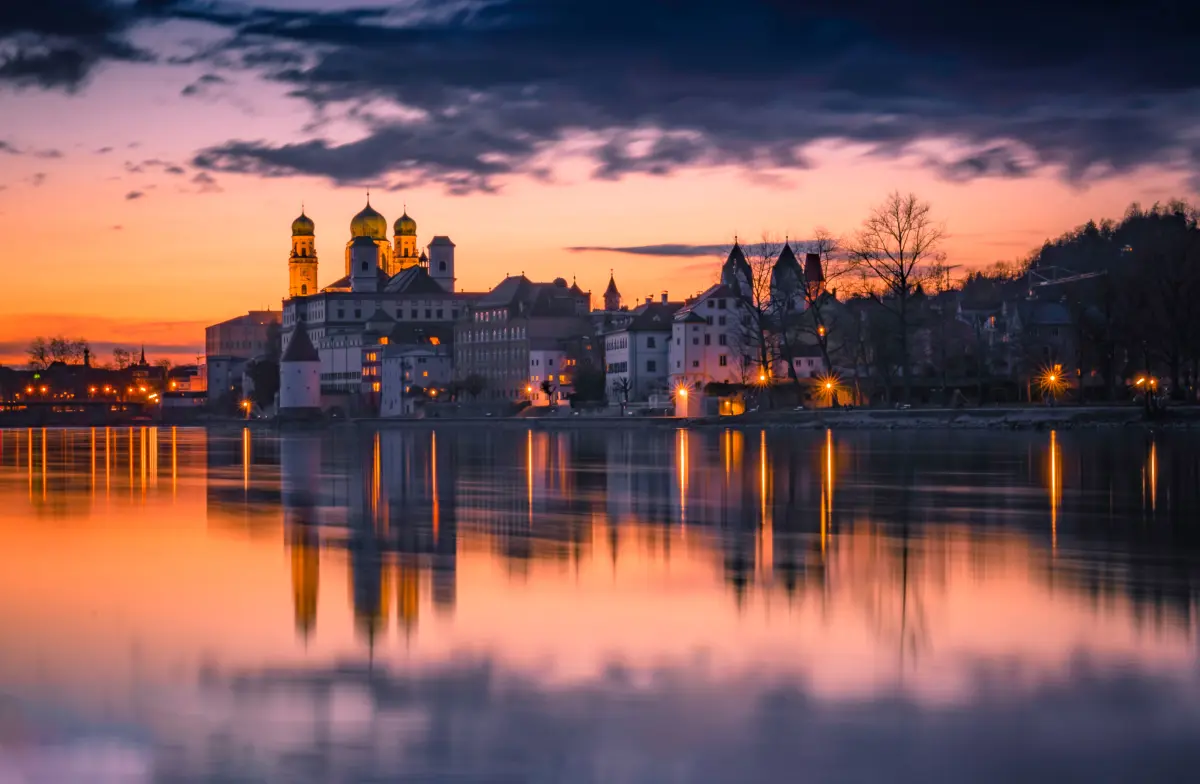 Die Stadt Passau mit ihrer Stadtsilhouette mit Kirchtürmen, die sich im ruhigen Fluss bei Sonnenuntergang unter einem dramatischen, farbenfrohen Himmel spiegeln.