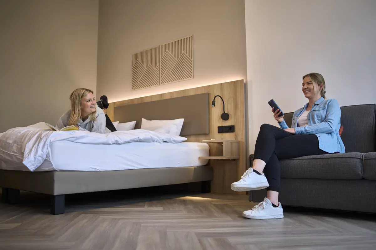 Zwei Frauen entspannen sich in einem modernen Hotelzimmer. Die eine liegt auf dem Bett und liest, die andere sitzt auf einem Sofa und telefoniert.