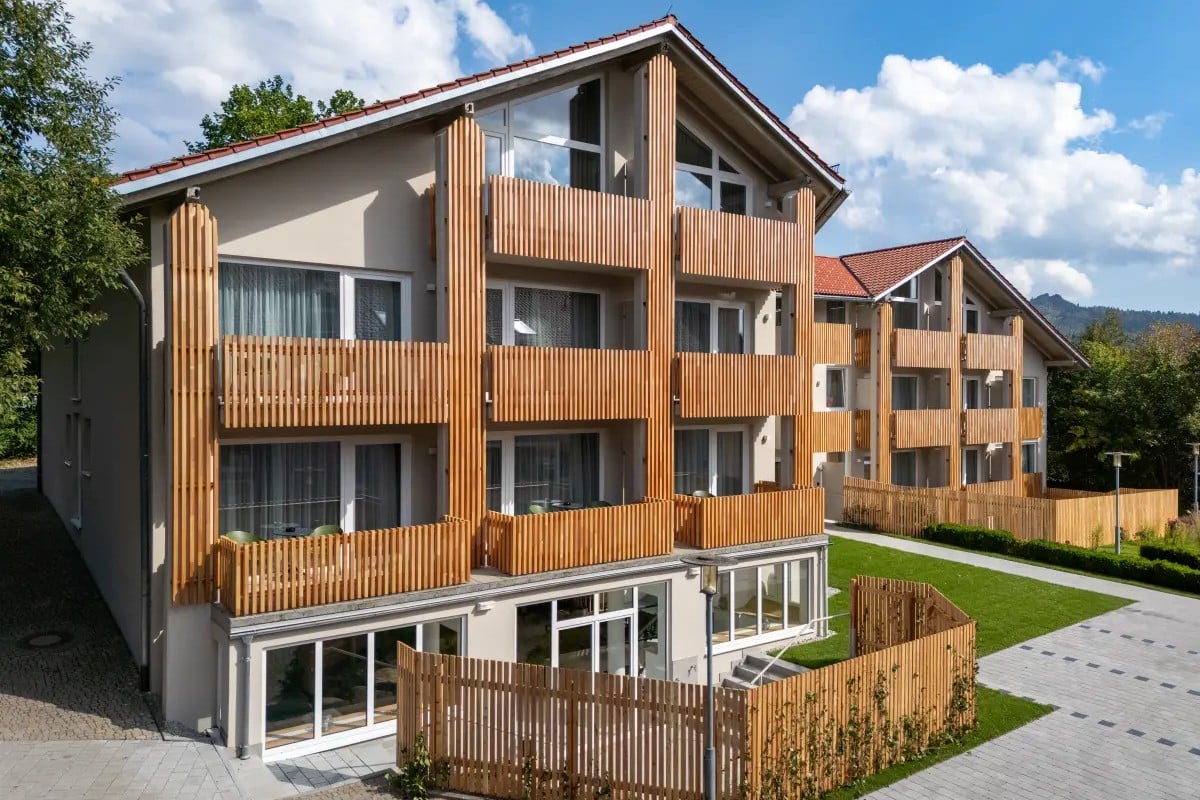 Modernes beigefarbenes Mehrfamilienhaus mit Holzbalkonen, großen Fenstern und einem gepflasterten Gehweg davor.