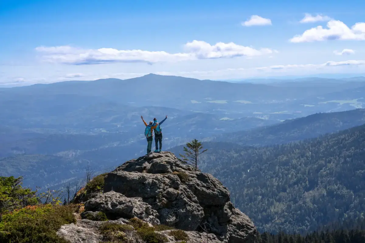 Zwei Wanderer stehen mit erhobenen Armen auf einem felsigen Gipfel im Bayerischen Wald und überblicken eine weite Berglandschaft unter blauem Himmel.